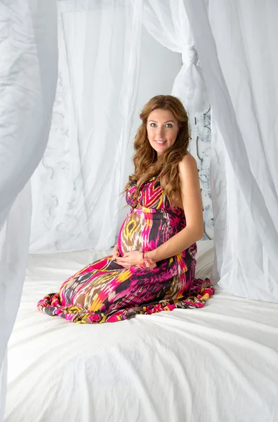 Young beautiful fashion pregnant woman in colorful dress sitting on a bed with a canopy on a white background