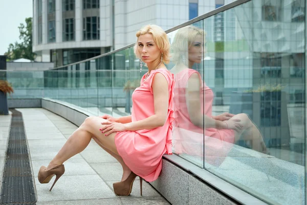 Outdoor summer fashion stunning portrait on pretty young blonde sexy woman dressed in pink dress wearing pastel colored jewelry on the background of the urban landscape.