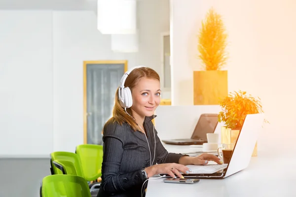 Young successful businesswoman in headphones with laptop computer surfing the internet at modern bright office. Woman using tablet computer and listening to the music during coffee break.