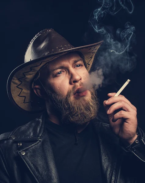 Smoking man with a beard and mustache wearing a cowboy hat.