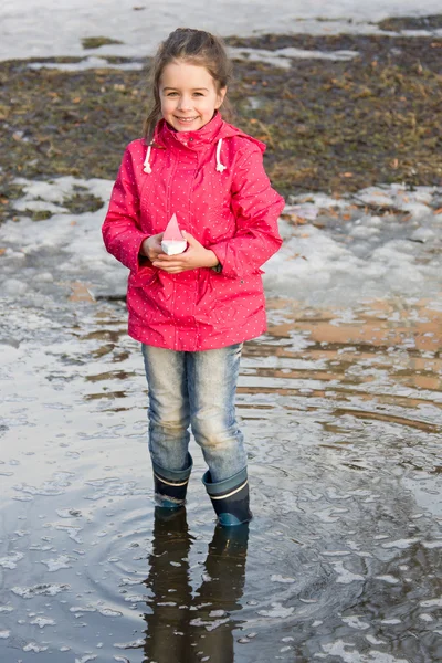 Happy little girl in rain boots playing with ships in the spring creek standing in water
