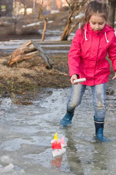 Cute little girl in rain boots playing with handmade ships in the spring creek standing in water