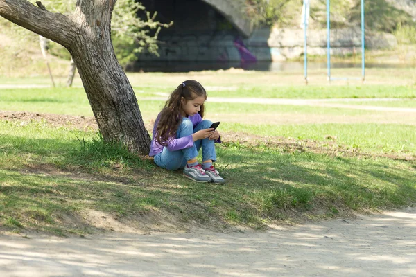 Cute little long-haired girl sitting on the grass with a mobile phone in her hands and stares at the phone's screen