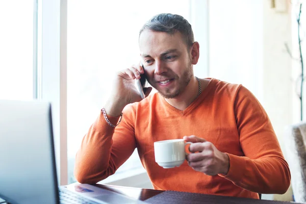 Handsome young man working with notebook, talking on the phone, smiling,  while enjoying coffee in cafe