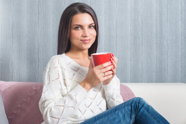 Woman  with a red mug in her hands, smiles sittings on the couch, sofa look at camera