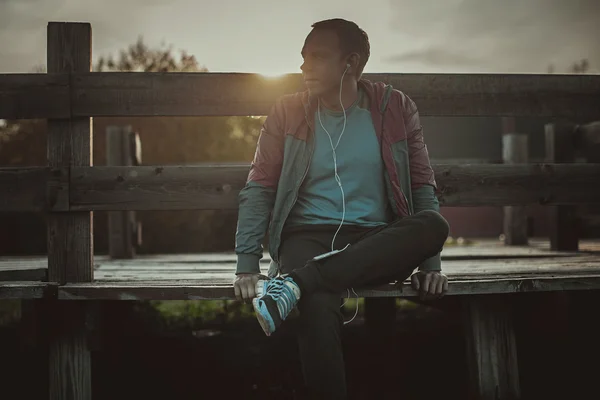 Tired runner sitting, relaxing and listening to music phone on a wooden pier, sport