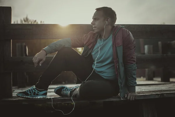 Tired runner sitting, relaxing and listening to music phone on a wooden pier, sport