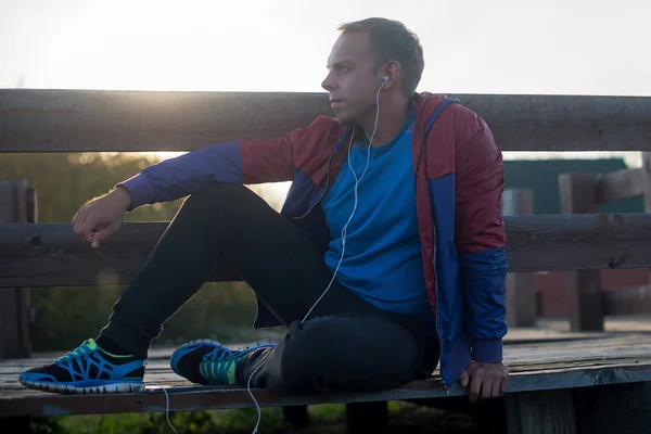 Tired runner sitting, relaxing and listening to music your phone on a wooden pier, sport.