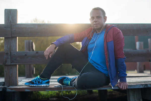 Tired runner sitting, relaxing and listening to music your phone on a wooden pier, sport.