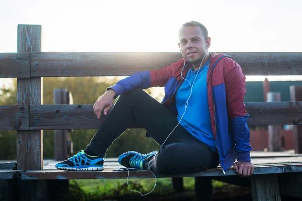 Tired runner sitting, relaxing and listening to music your phone on a wooden pier, sport.