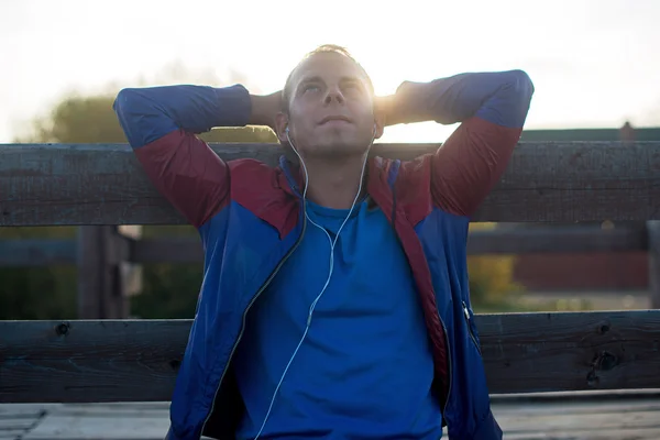 Tired runner sitting, relaxing and listening to music  your phone, eyes closed on a wooden pier, sport.
