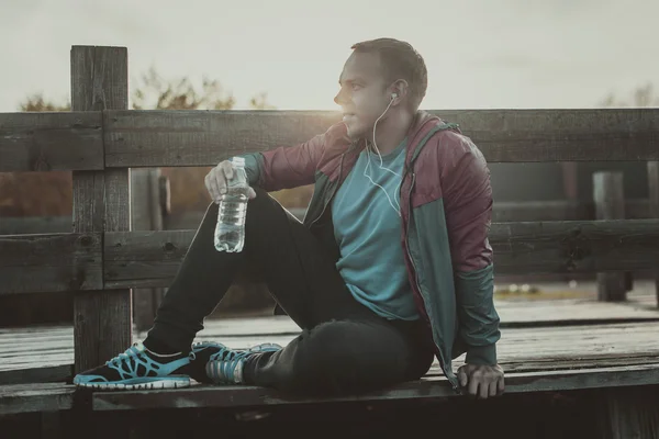 Tired runner sitting, relaxing and listening to music phone on a wooden pier, sport.