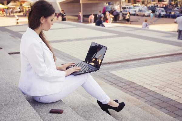 Beautiful brunette business woman in white suit with notebook on her lap, typing, working outdoors. Copy space