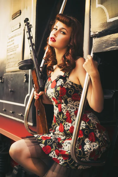 Beautiful sexy brunette retro woman with pin-up hairstyle and make-up in a shorts and shirt with a PCA gun in her hands on vintage train. Outdoors. Retro toned