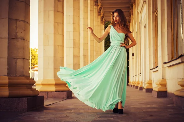 Beautiful blonde with a long curly hair in a long evening dress in motion outdoors near retro vintage building in summer sunset. Copy Space