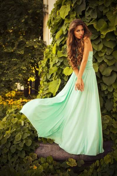 Beautiful blonde with a long curly hair in a long evening dress in motion outdoors near retro vintage building all in leaves in summer sunset