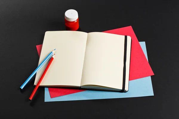 Open blank notepad with empty white pages laying with color paper and color pencils on black background. ideal for text