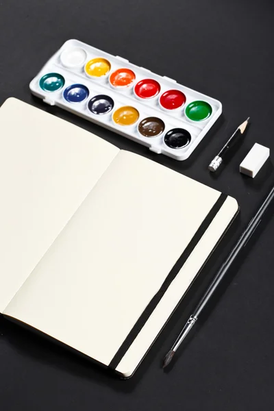 Open blank notepad with empty white pages laying with water-color palette brush pencil eraser on black background. ideal for text