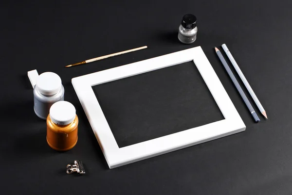 Black and white mock up with white wooden photo frame with black field and set of gold and silver gouache water-colors. Artist painter workplace