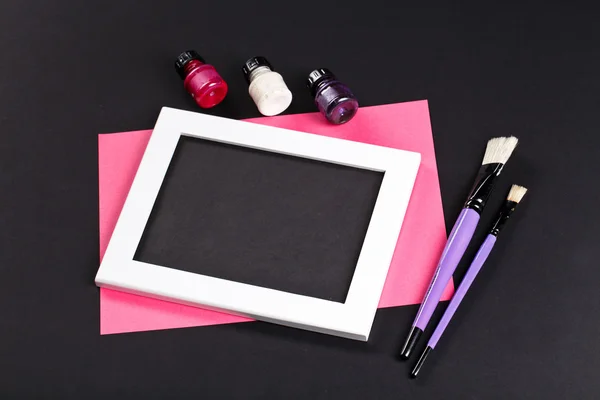 Black and pink template with white photo frame on pink paper with brushes and gouache. Painter workplace