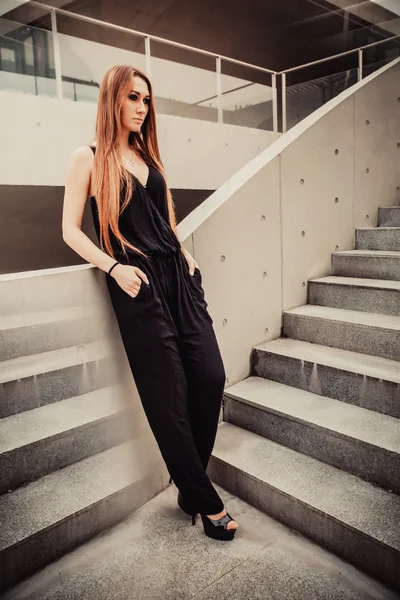 Red-haired girl model standing near the stairs in modern architectural environment.