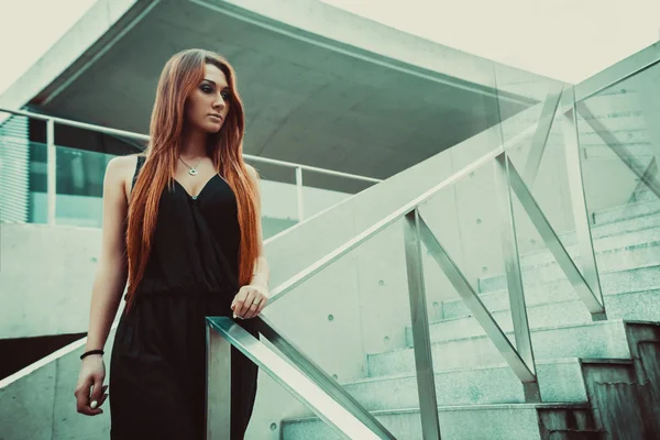 Red-haired girl model standing near the stairs in a modern architectural environment.