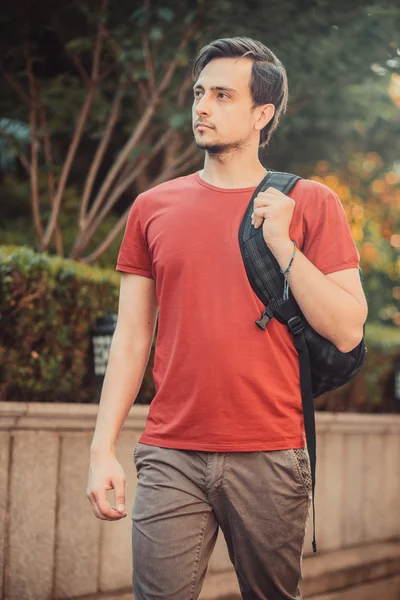 Man model in sport clothes and bag walks in Park near brick wall with bushes.