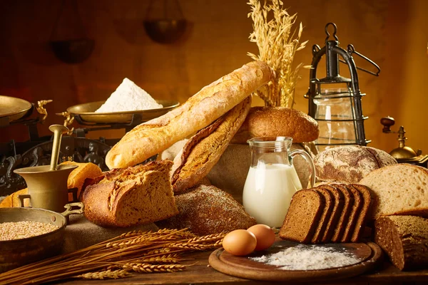 Still life with different kinds of bread, grain, flour on weight, ears of wheat, pitcher of milk and eggs