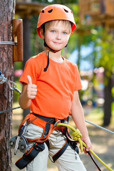 Cute boy shows thumb up with climbing equipment in an adventure park
