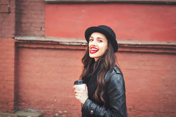 Trendy woman in hat  with drink outdoor. Young  woman with red lips  in the street and holding cup of coffee