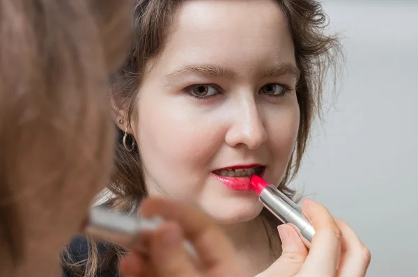 Young woman is looking in mirror and applying red glossy lipstick oh her lips.