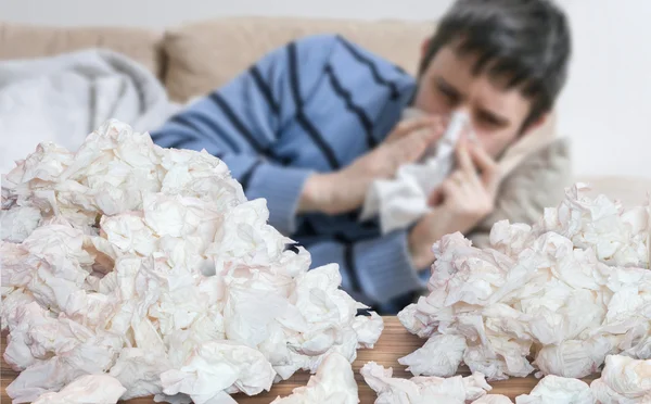 Funny sick man who has flu or cold is blowing his nose. Pile of tissues in front.