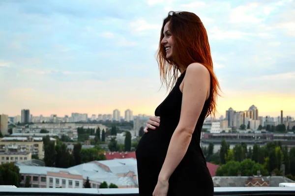 Young beautiful and happy pregnant redhaired girl over the view of sunset and the city looking forward of future motherhood and expecting her baby in a romantic black dress