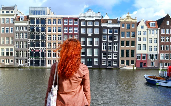 Redheaded woman in leather jacket standing with her back to the camera and looking at traditional buildings of Amsterdam city near the canal and boats