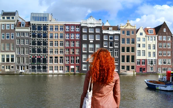 Redheaded woman in leather jacket standing with her back to the camera and looking at traditional buildings of Amsterdam city near the canal and boats