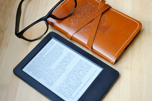 Black ereader with reading glasses and an old notebook in leather cover on wooden table