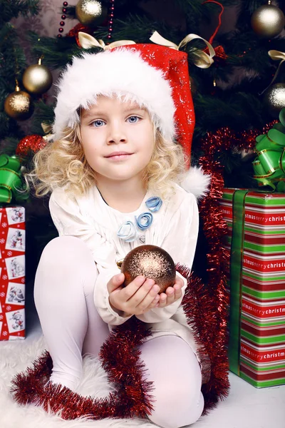 Cute little girl in Santa's hat sitting beside a Christmas tree