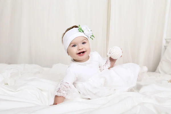 Cute little baby girl in white clothes, sitting on bed, playing with toy