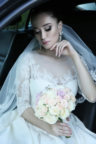Bride with dark hair in luxurious wedding dress with  bouquet of flowers sitting in car