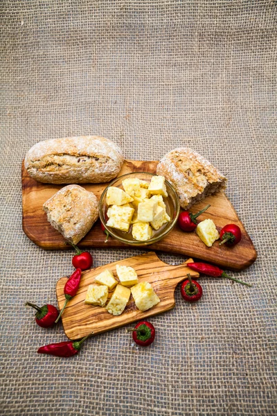 Composition with olive wood, cheese pieces in olive oil, bread and hot peppers with burlap texture