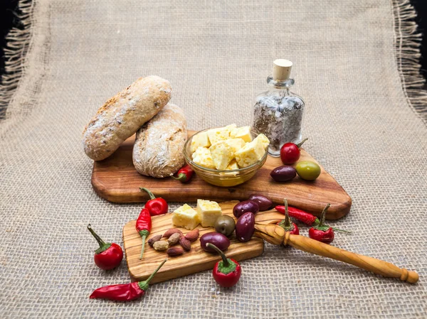 Composition with olive wood, olives, cheese pieces in olive oil, bread and spices with burlap texture in the background