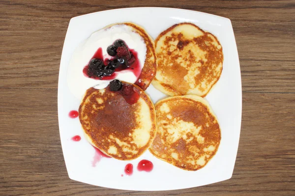 Pancakes with sour cream and berries in a on a square white plate. The cheesecakes on the plate drizzled with jam and sour cream. Hot cakes for Breakfast in white plate on wooden texture, top view