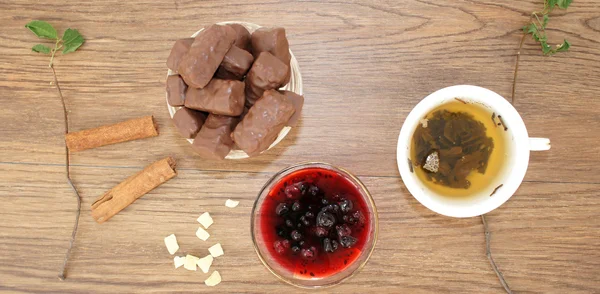 A mug of tea, chocolates and a vase with currant jam on wooden background. Mug of green leaf tea, a bowl of candy and jam, top view. A mug of tea, a bowl of candy, jam, cinnamon and candied fruit.