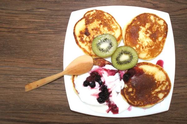 Pancakes with sour cream, berries and kiwi in a on a square white plate with wooden spoon.Hot pancakes in white plate on wooden texture, top view