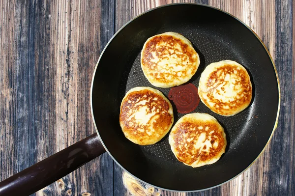 Pancakes in a frying pan on a wooden texture. Frying pan with hot cheesecakes on a wooden texture. Just cooked pancakes in the pan, top view. Fried crispy hot pancakes in a frying pan.