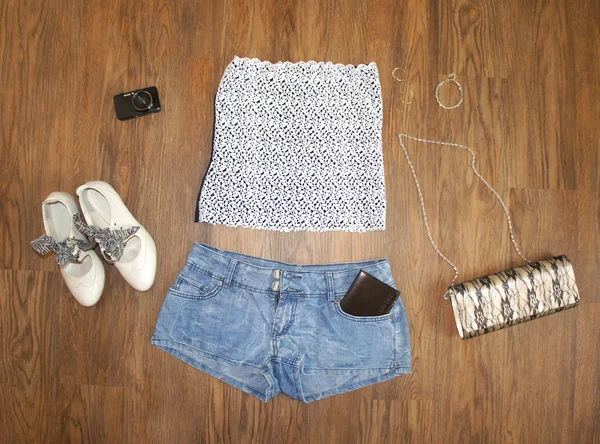 Flat lay feminine clothes and accessories collage: lace top, denim shorts with a passport, clutch bag, shoes, camera and decorations on wooden background