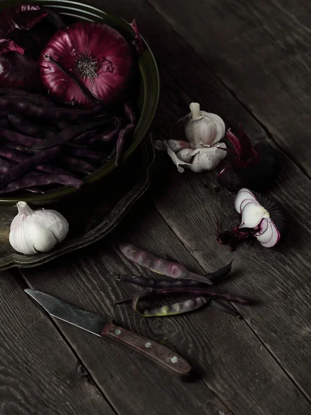 On a dark wooden table in green clay bowl vintage metal platter placed vegetables, blue onion, beans, garlic, old knife.