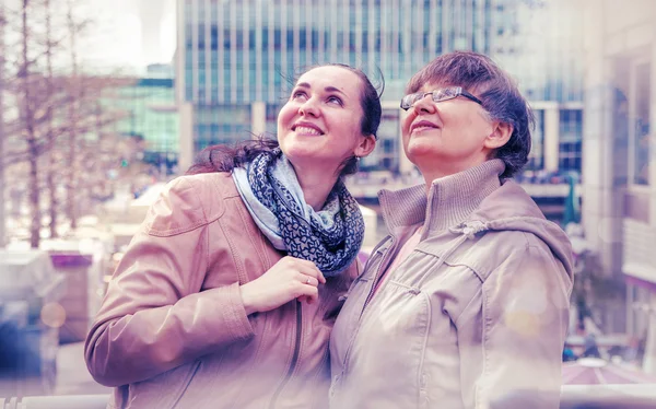 Outdoor family portrait of pension age Mother and her daughter in the city, smiling and looking around. Two generation, happiness and care concept