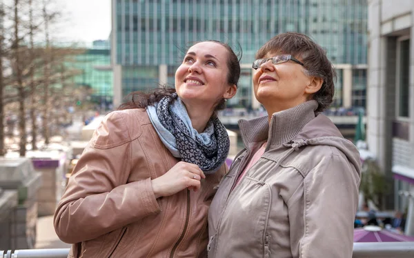 Outdoor family portrait of pension age Mother and her daughter in the city, smiling and looking around. Two generation, happiness and care concept