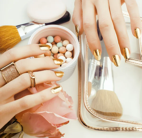 Woman hands with golden manicure and many rings holding brushes, makeup artist stuff stylish, pure close up pink flower rose among cosmetic